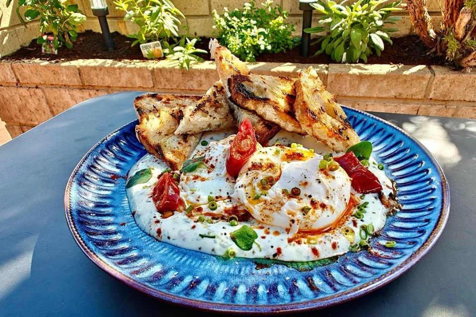 A blue ceramic plate with Turkish eggs topped with sliced tomatoes, green herbs, and a fried egg, alongside grilled bread pieces, set outdoors on a patio with plants in the background.