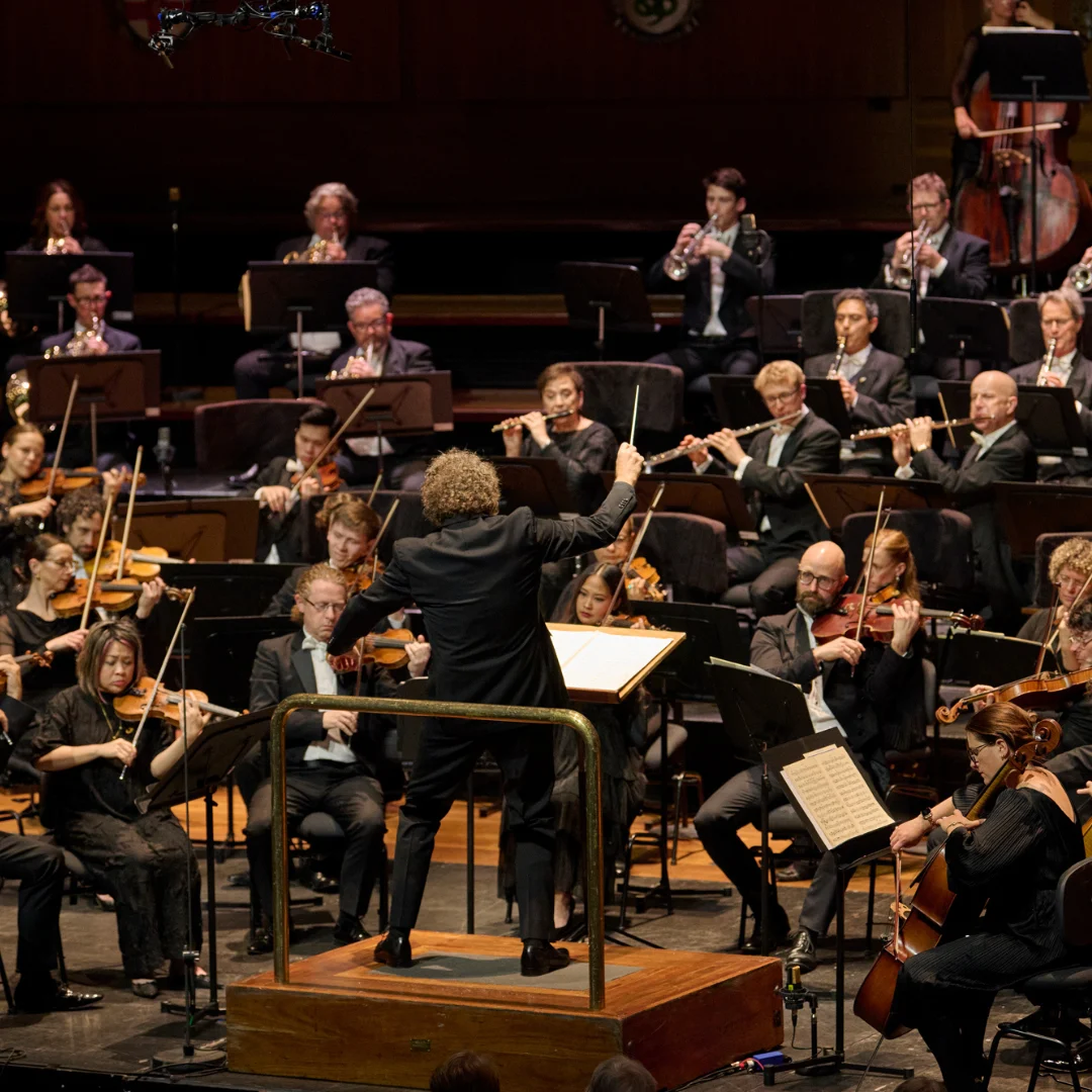 An orchestra performs on stage, with a conductor standing on a podium leading musicians playing various instruments, including violins, cellos, flutes, and brass, in a formal concert setting.