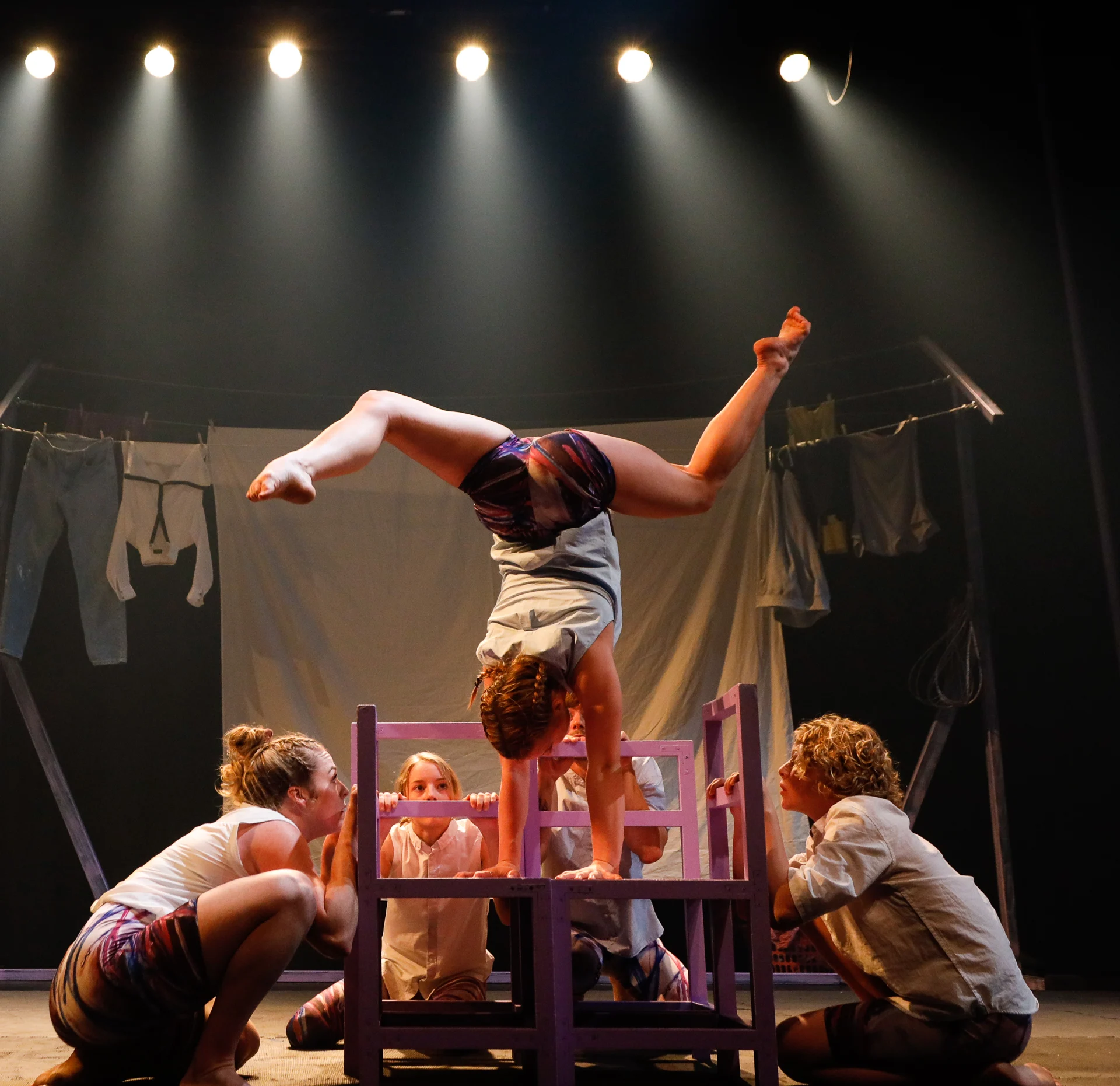 Four people watch as a performer does a handstand on a purple structure on stage, with dramatic lighting and hanging clothes in the background.