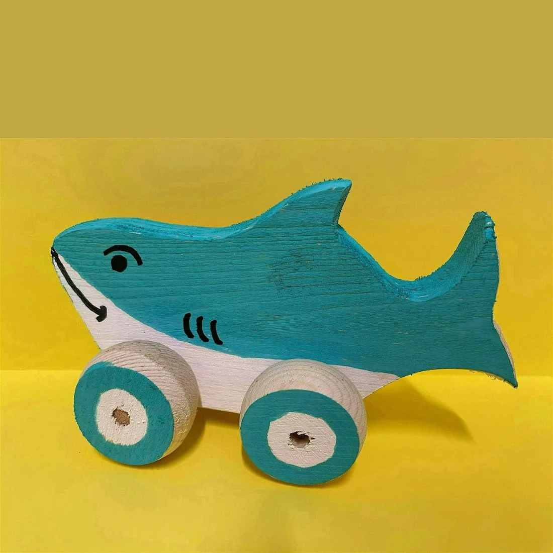 A teal wooden toy shark on a yellow background, with black line details on its body and two large round wheels painted teal and white.