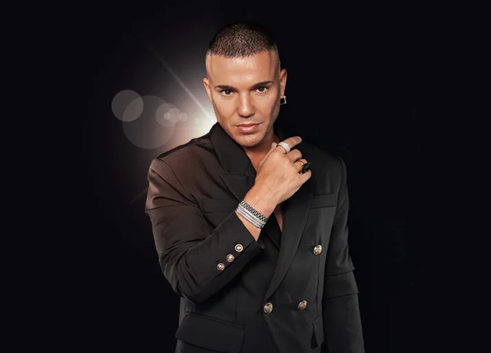 A man with short hair wearing a dark double-breasted blazer and bracelets, posing against a black background.