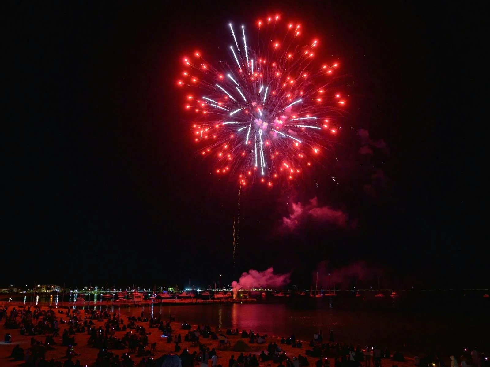 Large red fireworks bursting above a beach filled with spectators