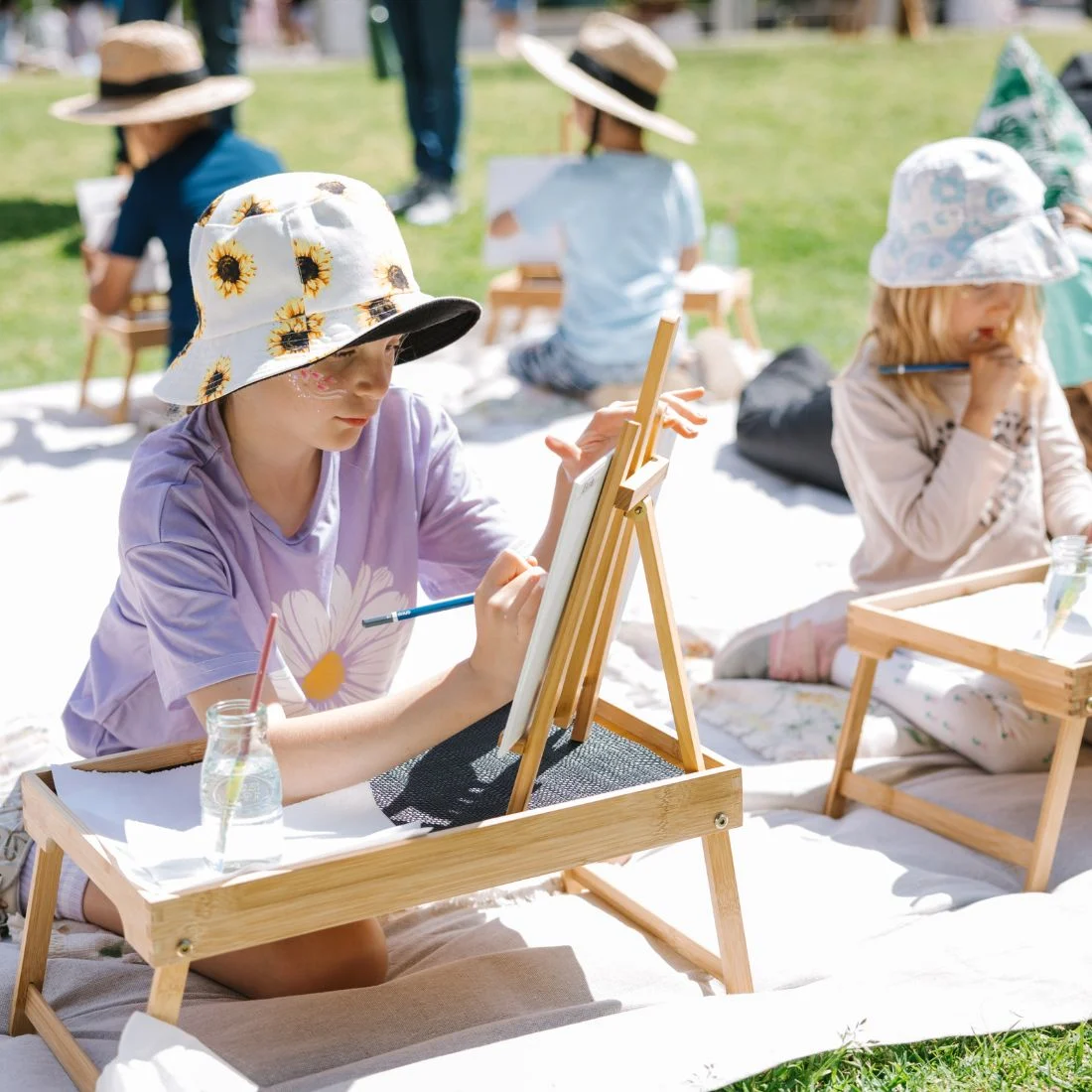 Children sit outdoors on blankets, wearing sun hats, and paint on canvases set on small wooden easels. Sunlight shines on the grassy area as they focus on their artwork, drinks in glass jars beside them.