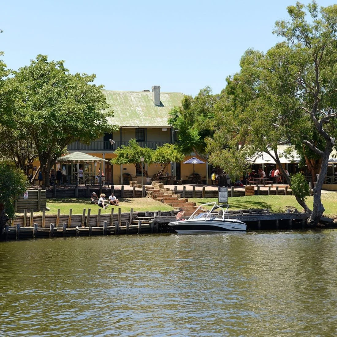 A small motorboat is docked by a wooden pier in front of a rustic, two-story building surrounded by trees. People are sitting outside on a sunny day near the waters edge.