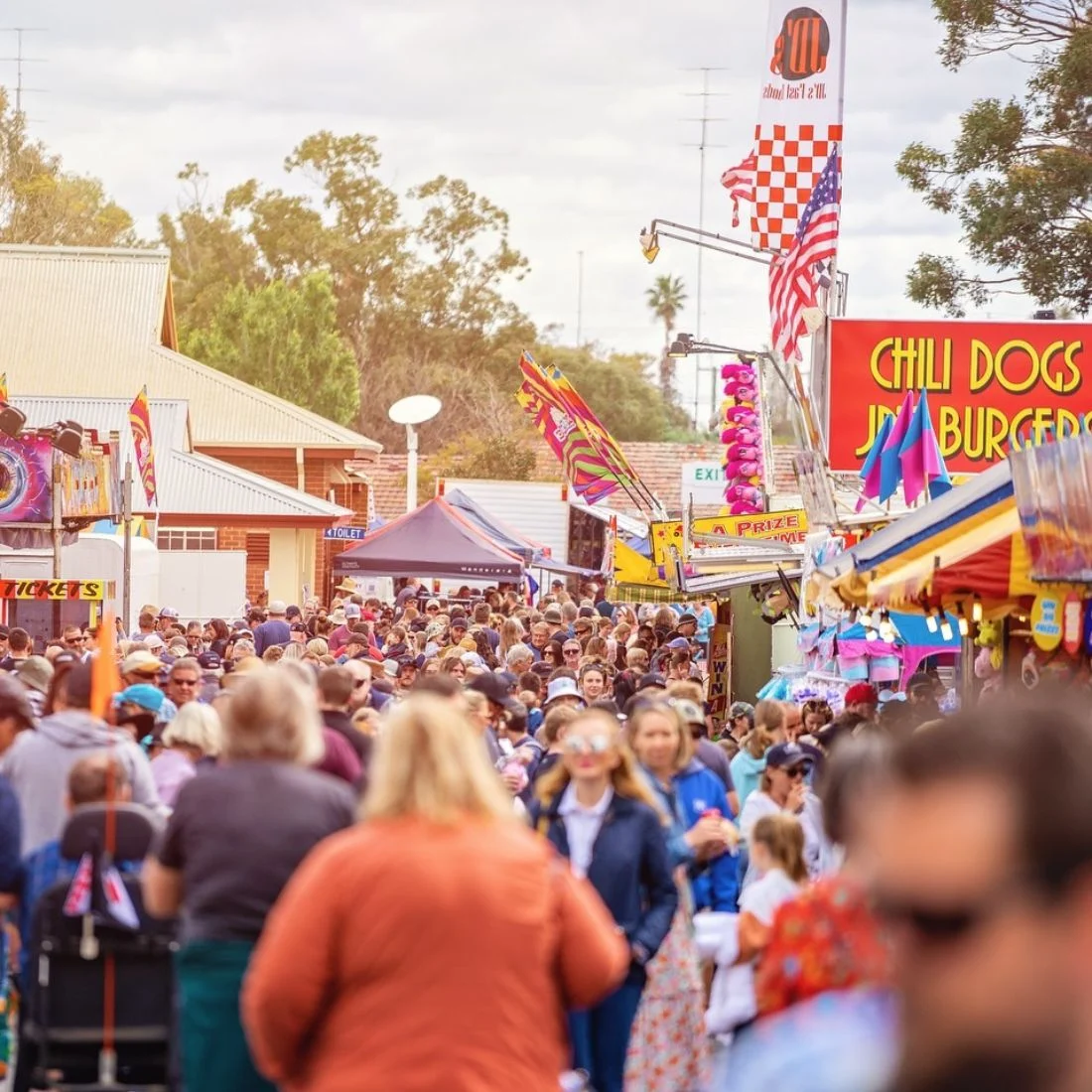 A large crowd walks through a lively fair with colorful food stalls, including signs for chili dogs and burgers, under a partly cloudy sky.