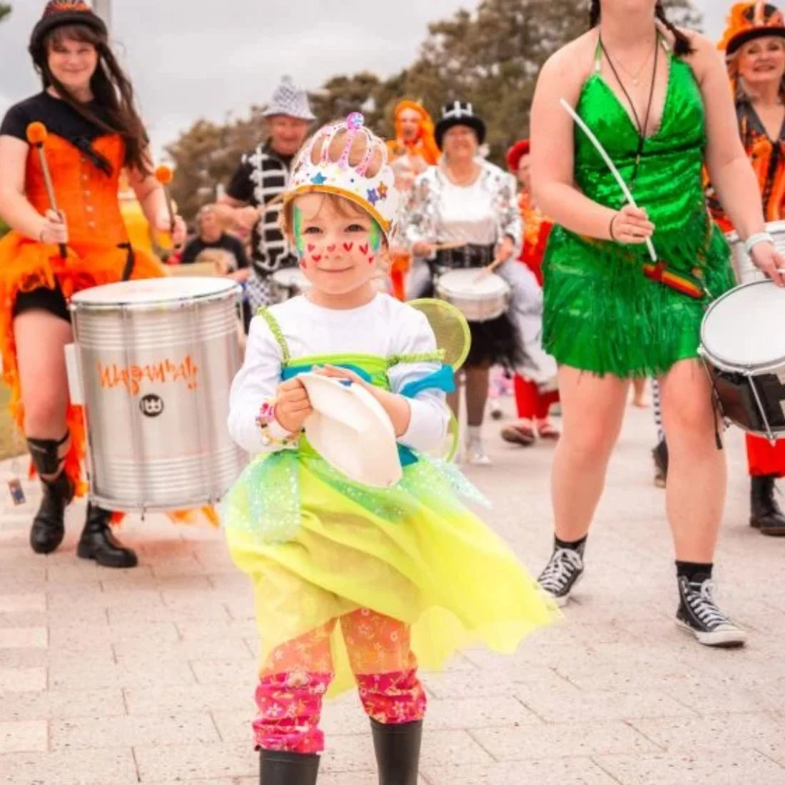 A child in a colorful costume and crown stands in front of a festive parade, with adults in bright outfits playing drums and marching behind them on a paved street.