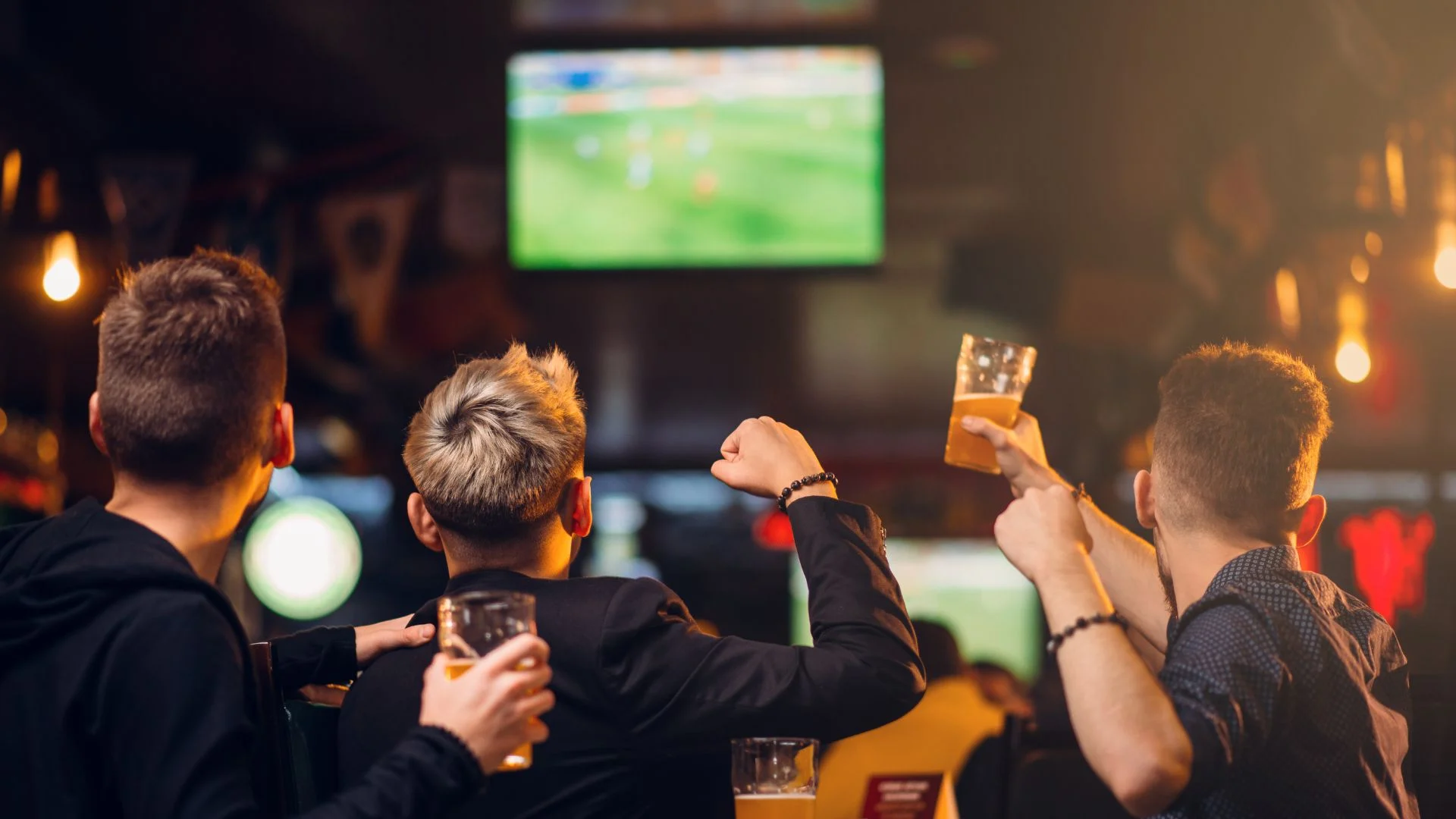Three people in a bar watch a soccer game on a TV screen, holding drinks and raising their arms in excitement.