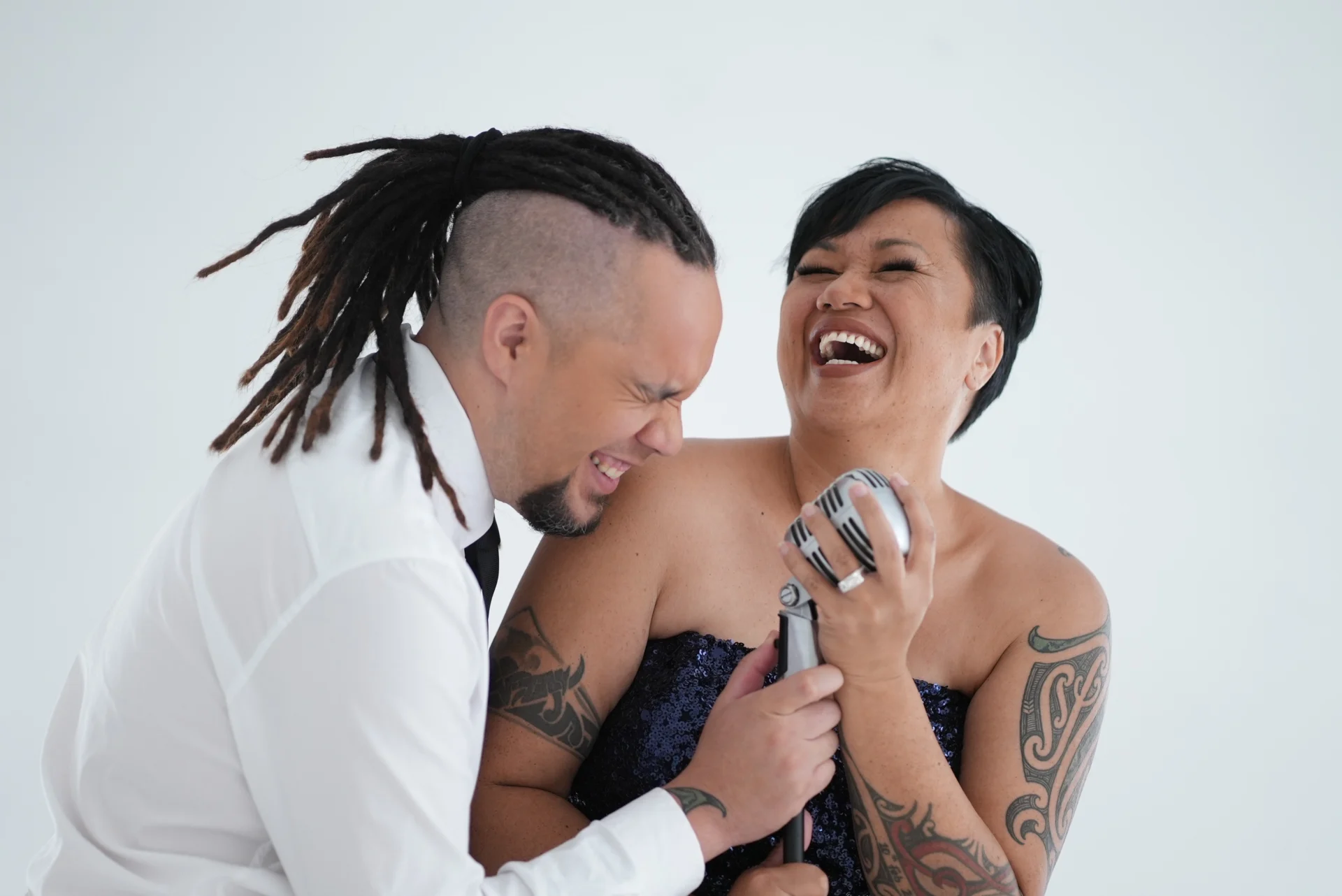 Two people laughing together, one holding a vintage microphone. Both have tattoos, and the person on the left has dreadlocks and wears a white shirt, while the person on the right has a strapless top.