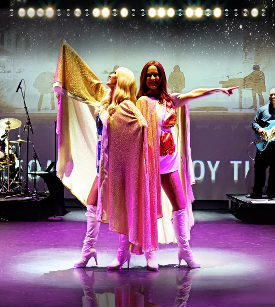 Two performers wearing sparkly capes and white boots strike a pose on a stage, backed by a band.