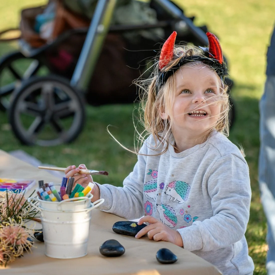A young girl wearing devil horns headband smiles while painting rocks at a table.