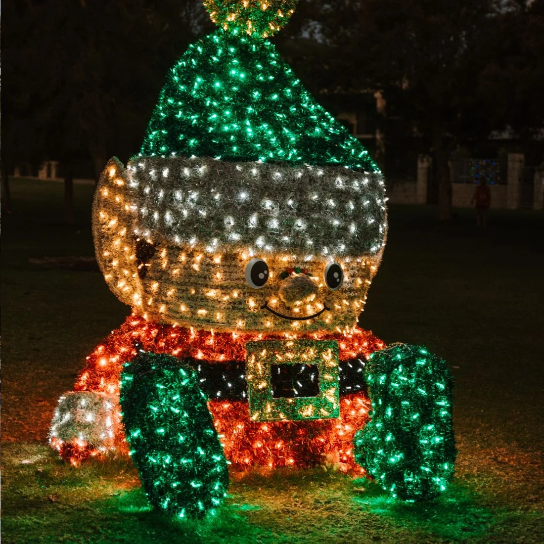 A large outdoor light display in the shape of a sitting elf wearing a green hat and red tunic.
