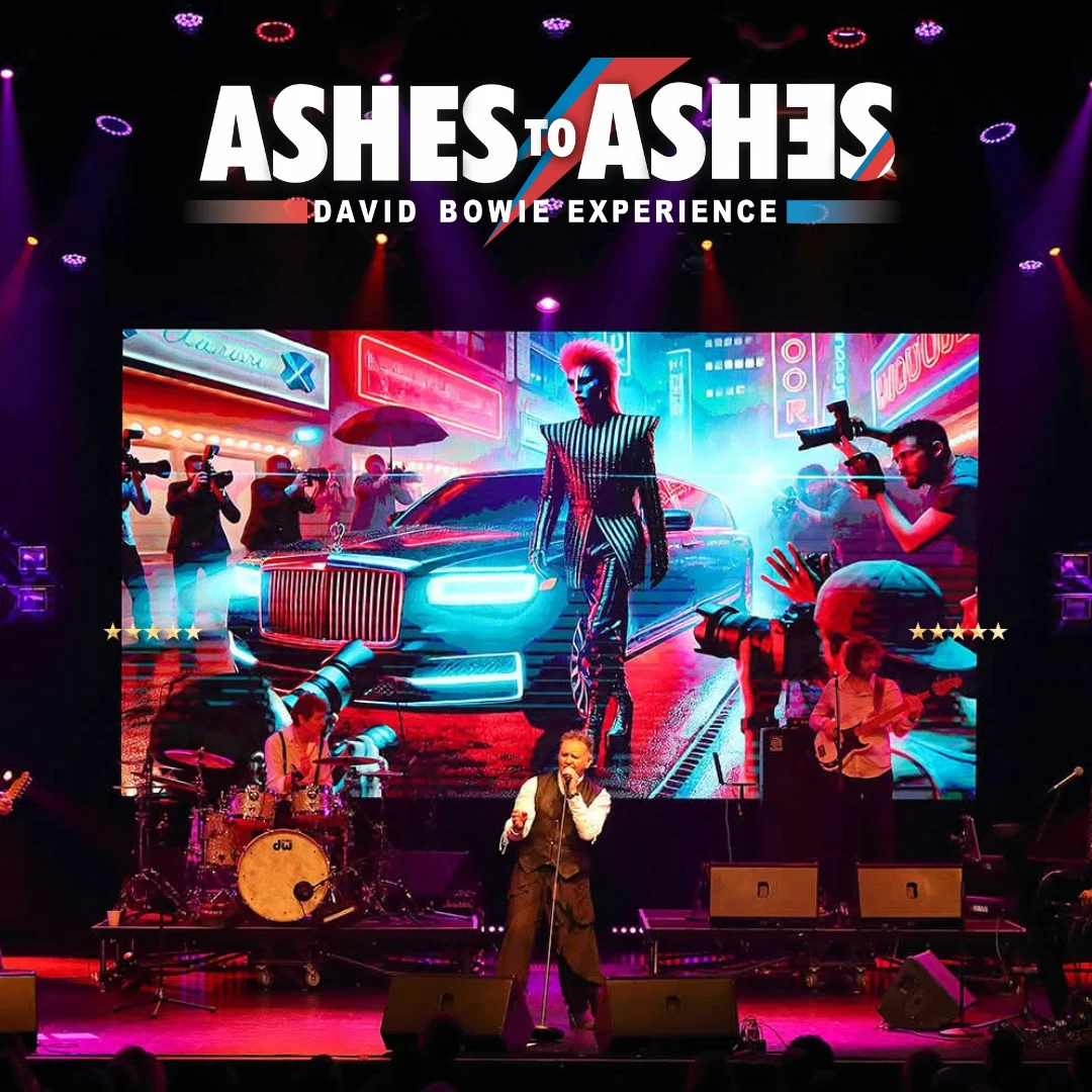 A David Bowie tribute band performs on a stage in front of a large screen displaying a stylized image of Ziggy Stardust next to a car and photographers.