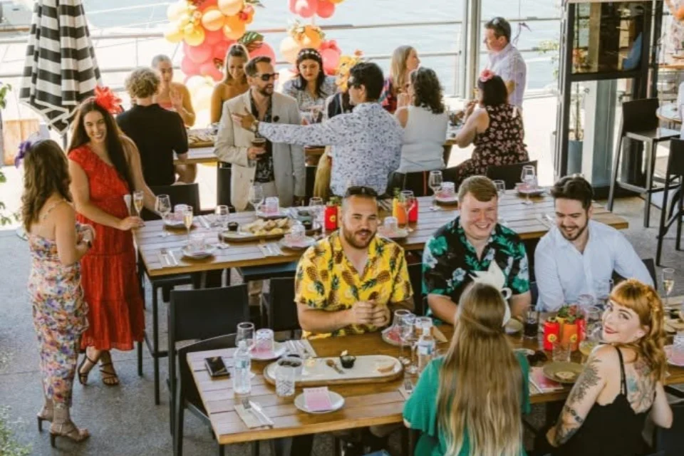 People chatting and smiling at a long outdoor banquet table during a birthday celebration, with colourful balloon and flower decorations overhead and children standing nearby, drinks and plates set out.