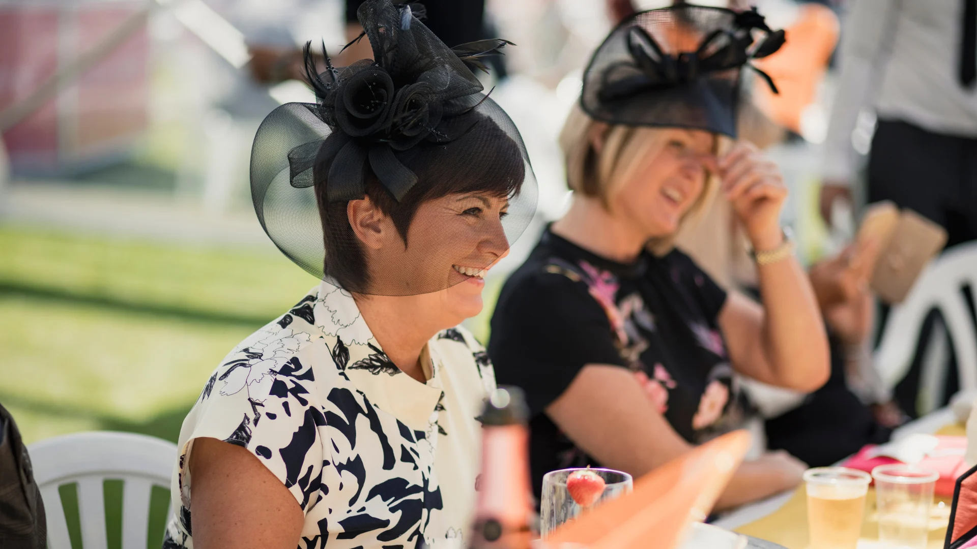 A woman wearing a black fascinator and white patterned dress smiles while seated outdoors at a table with red and clear drinks; a seated person in a dark hat and others sit in the background.