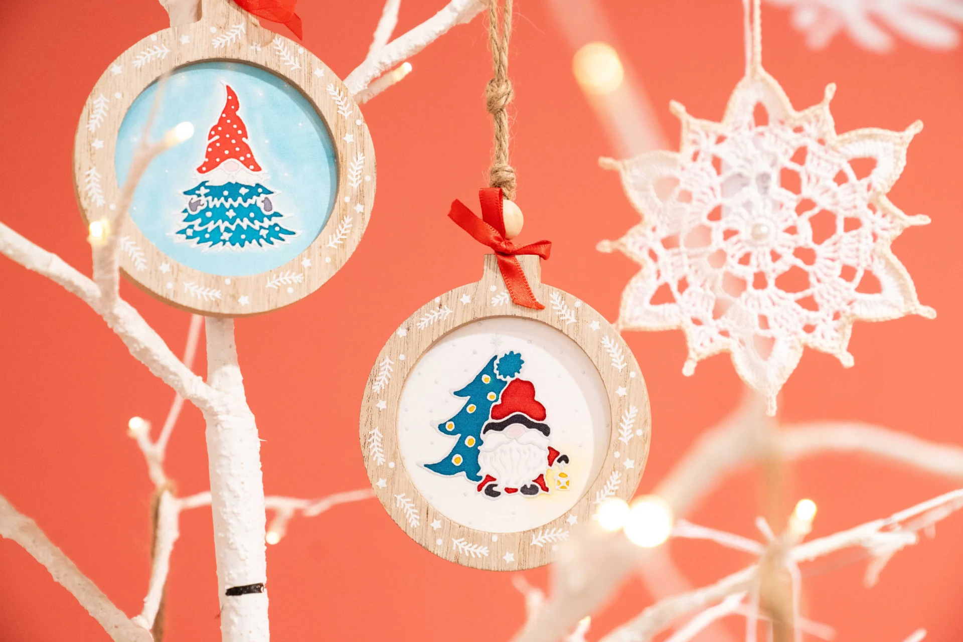 Two round wooden ornaments featuring gnome and Christmas tree illustrations hanging alongside a white crocheted snowflake ornament on a white decorative branch.